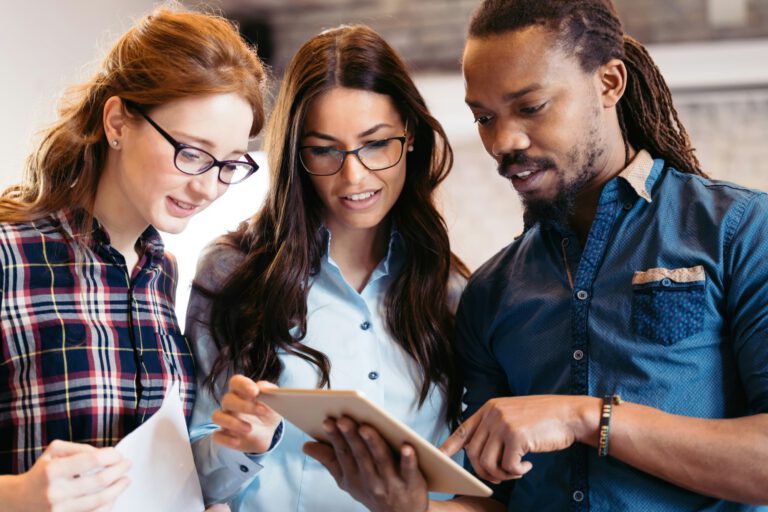 Three people standing together, discussing web design Carlisle projects as they review information on a digital tablet, with one person holding papers.