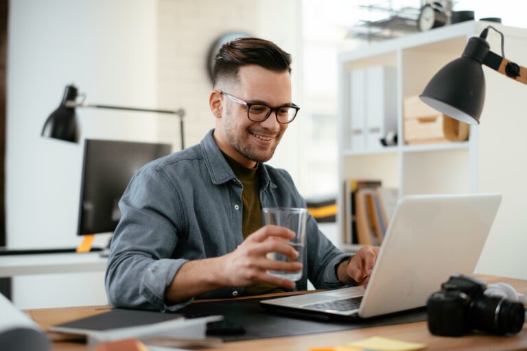 A smiling man with glasses sits at a desk, holding a glass of water while typing on his laptop, perhaps working on a Carlisle web design project. The bright, modern office features shelves, a monitor, camera, notebook, and lamp.